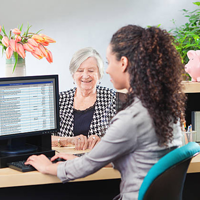 A senior woman bank customer making a financial transaction with a bank teller over the counter in a retail bank. The young woman bank teller is working on her computer terminal providing the customer service. Photographed from behind the shoulder of the bank teller in a horizontal format.